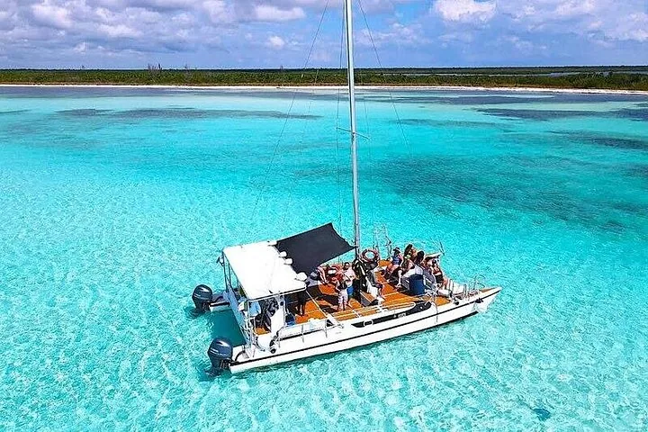 Luftaufnahme eines weißen Katamarans, der in flachem türkisfarbenem Wasser vor Anker liegt, mit Personen an Bord.
